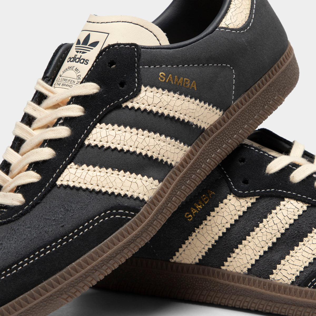 adidas Juniors' Samba OG. Close-up view of tongue and lacing highlighting carbon dark grey leather construction, cracked leather warm vanilla serrated 3-Stripes, gold foil "Samba" logo text, warm vanilla laces, and warm vanilla leather tongue with dark grey adidas Trefoil logo print. 