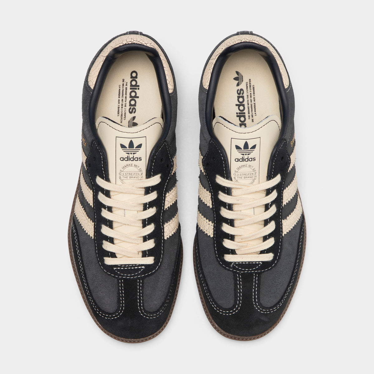 adidas Juniors' Samba OG. Top-down overhead view of a pair of low-top sneakers displaying carbon dark grey suede T-toe overlays, carbon suede uppers, warm vanilla laces, warm vanilla leather tongues with dark Trefoil logos, and off-white insoles with dark grey adidas Trefoil branding. 