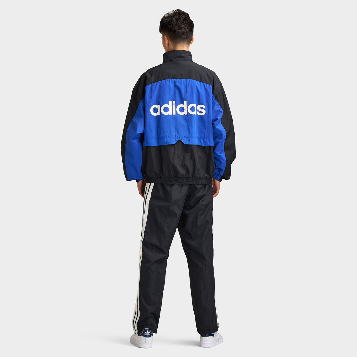 adidas Munich 93 Tracktop. Full-body back view of model wearing blue and black track jacket with large white adidas branding and matching black pants.