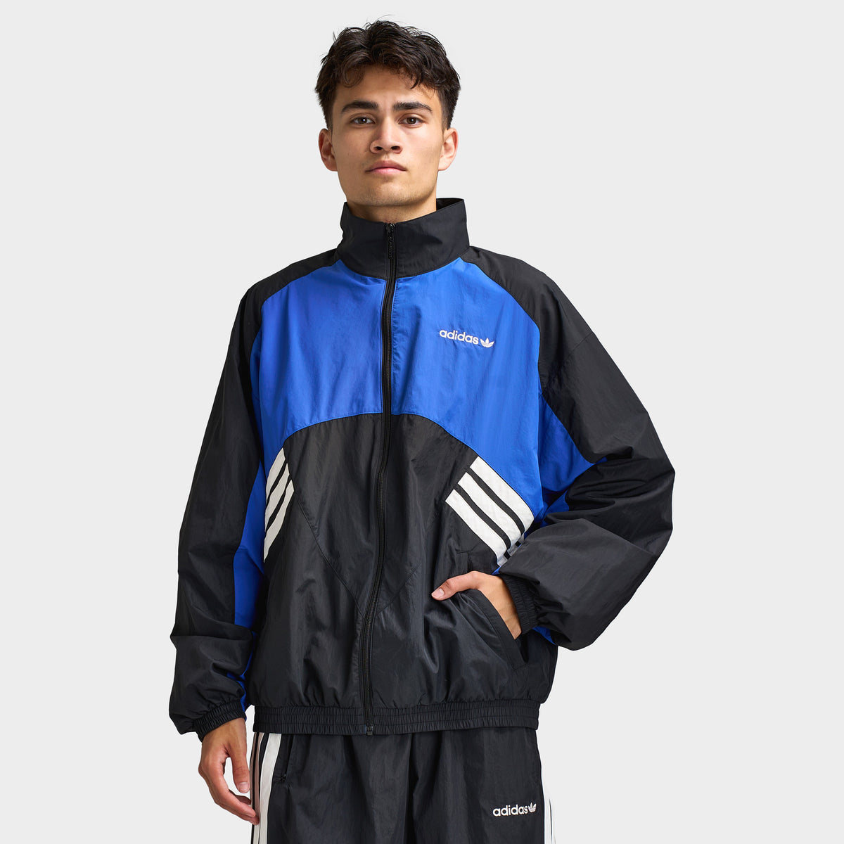 adidas Munich 93 Tracktop. Front view of model wearing blue and black track jacket with high collar, full-zip closure and contrast white adidas branding.