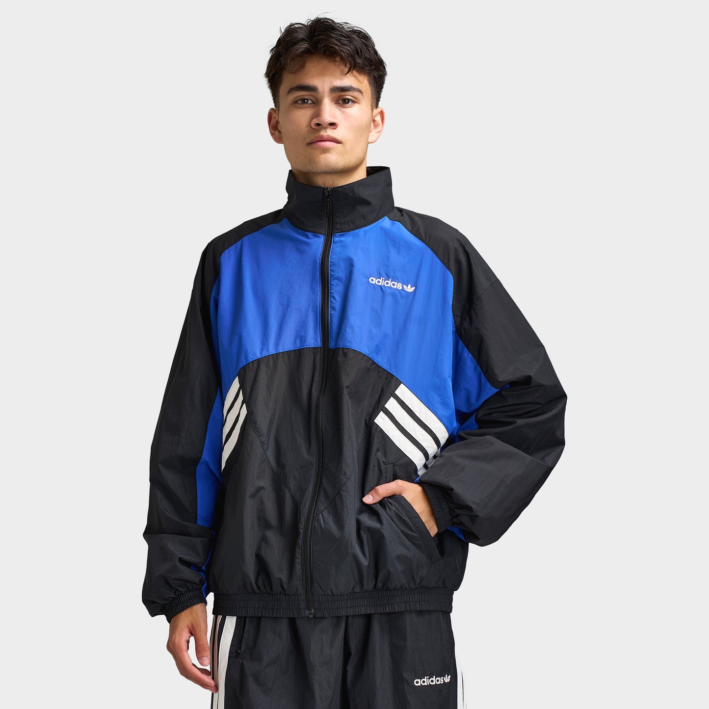 adidas Munich 93 Tracktop. Front view of model wearing blue and black track jacket with high collar, full-zip closure and contrast white adidas branding.