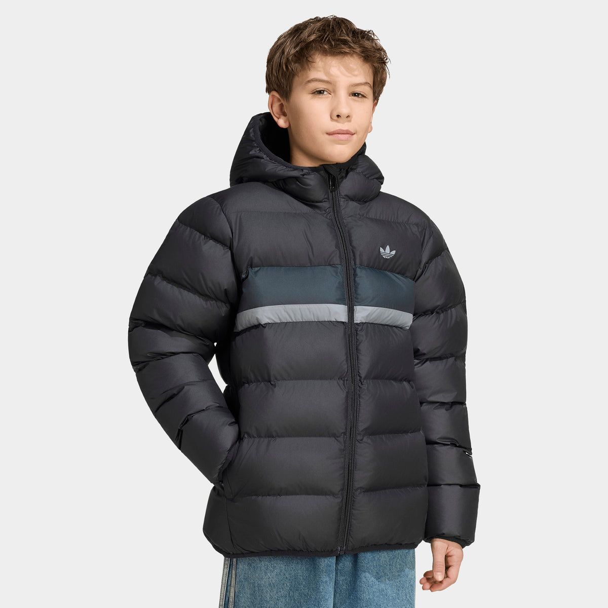 adidas Juniors' Blocking Regular Puffer. Three-quarter front view of a black padded puffer jacket worn by a youth model, featuring horizontal quilted baffle construction, charcoal and silver chest color-blocking stripe, white adidas Trefoil logo on the left chest, full-zip closure, attached hood, and elasticated cuffs. 