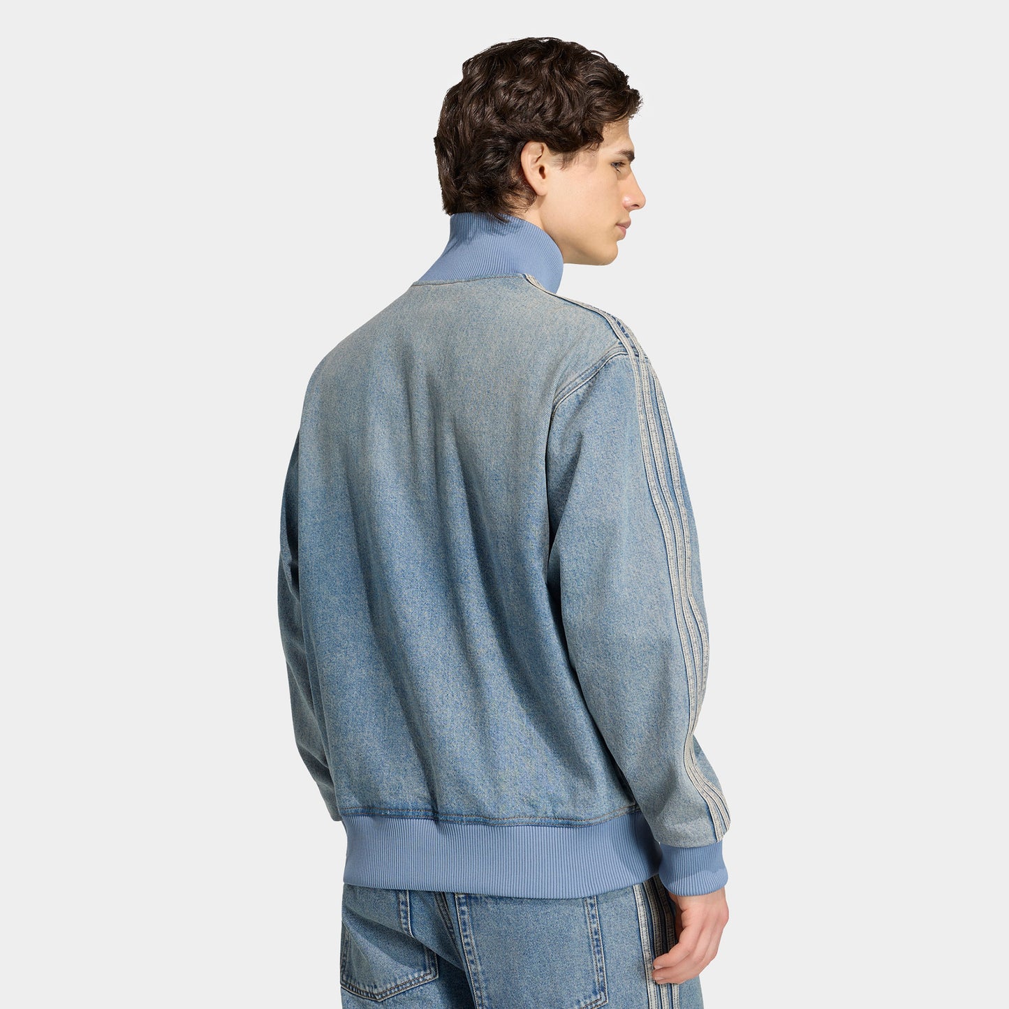 adidas Firebird Adicolor Track Top. Back view of model wearing worn blue denim track jacket with plain back, high collar and striped sleeves.