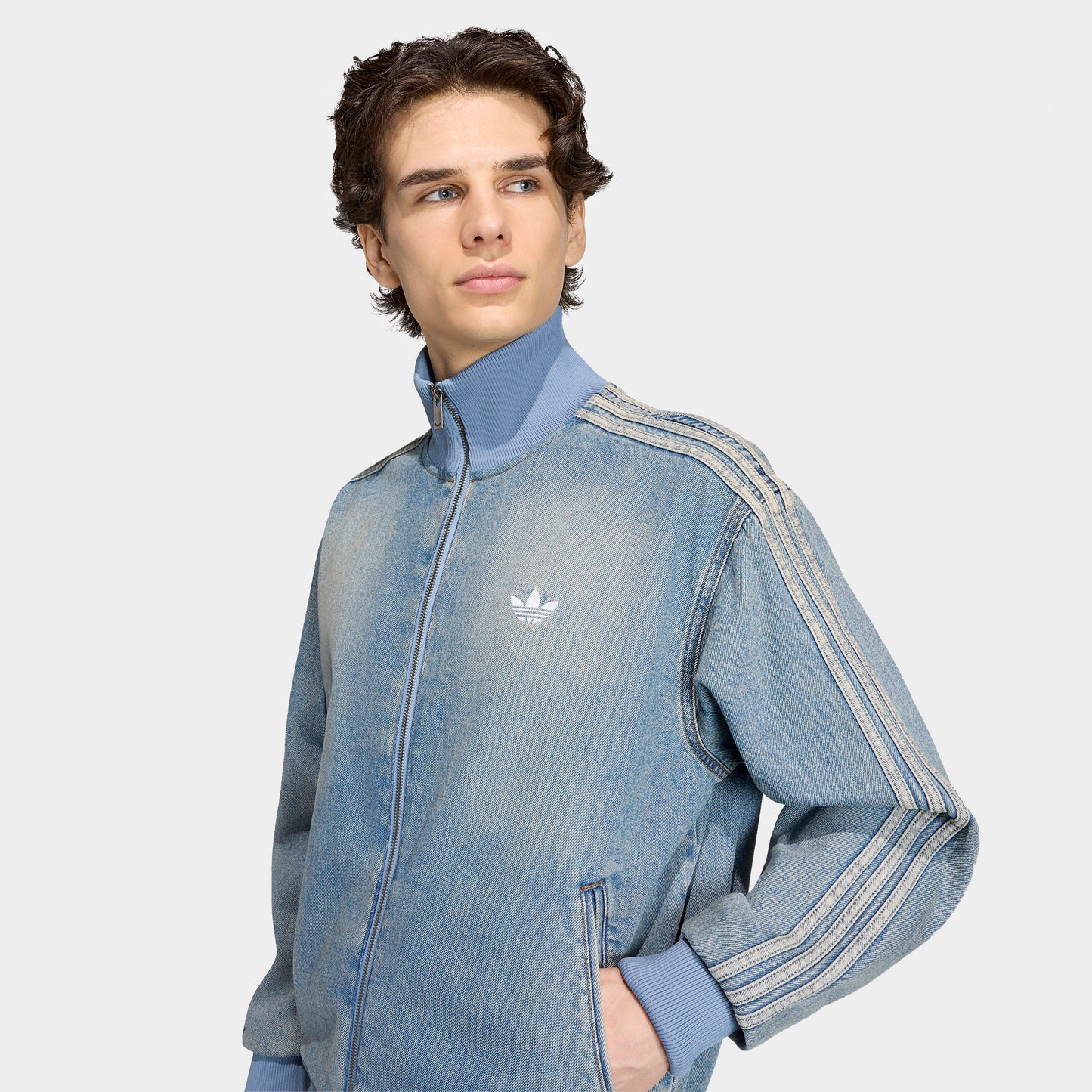 adidas Firebird Adicolor Track Top. Three-quarter view of model wearing worn blue denim track jacket with full-zip closure, high collar and white Trefoil logo.