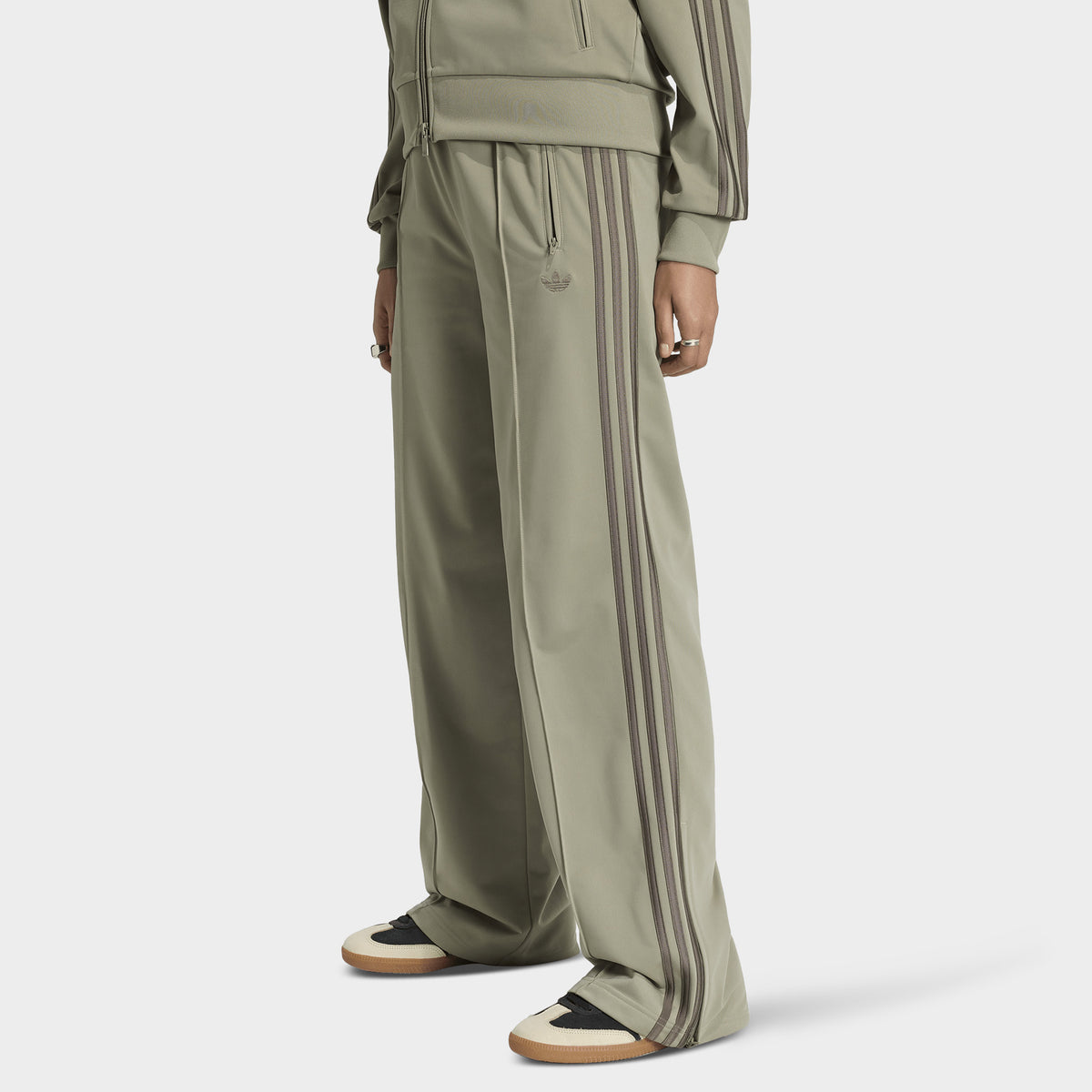 adidas Firebird Loose Track Pants. Front view of model wearing silver-pebble branch track pants with wide legs, elastic waistband and tonal side stripes.