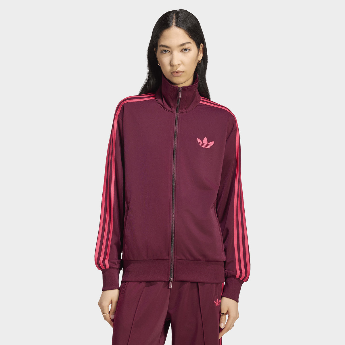 adidas Firebird Track Jacket Loose. Front view of model wearing a maroon track jacket with power pink 3-Stripes, a loose fit, a full-zip fastening and an embroidered Trefoil logo at the chest.