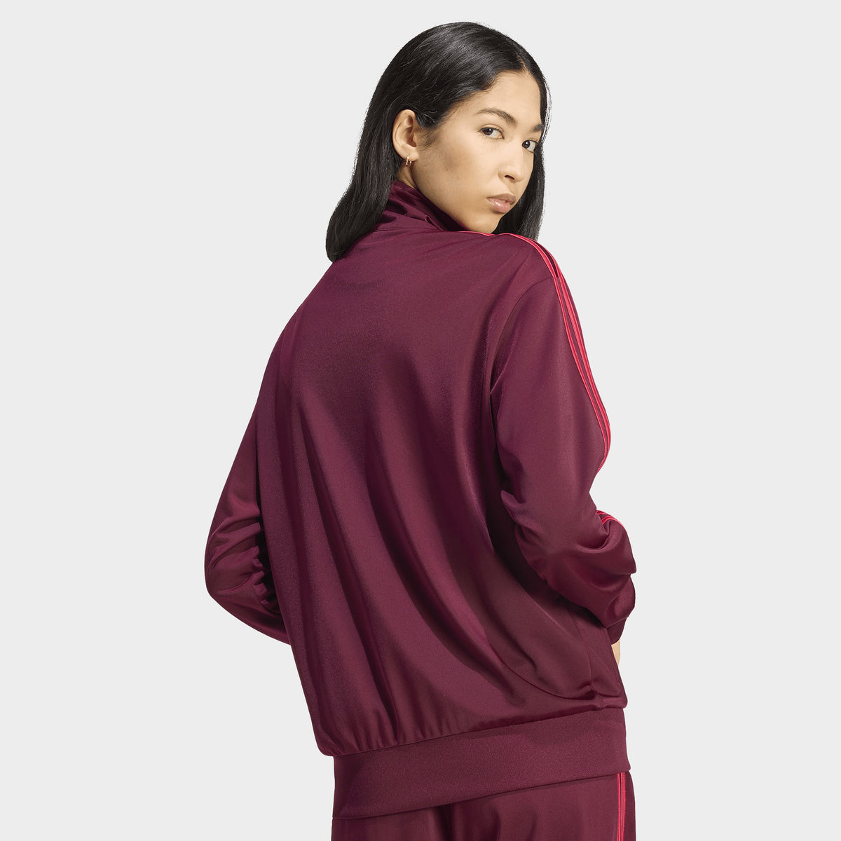 adidas Firebird Track Jacket Loose. Back view of model wearing a maroon track jacket with power pink 3-Stripes and a loose, relaxed fit.