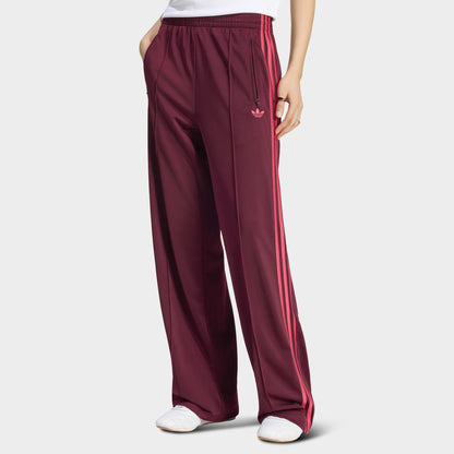 adidas Firebird Loose Track Pant. Front view of model wearing maroon track pants with power pink side stripes, a loose fit and an embroidered Trefoil logo at the hip.