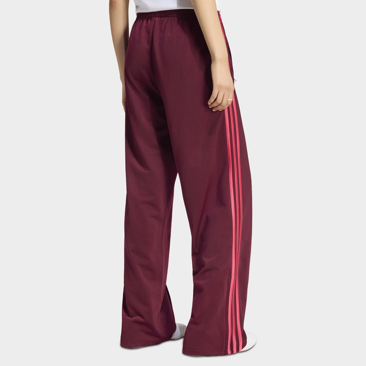 adidas Firebird Loose Track Pant. Side view of model wearing maroon track pants with power pink side stripes and a relaxed wide-leg fit.