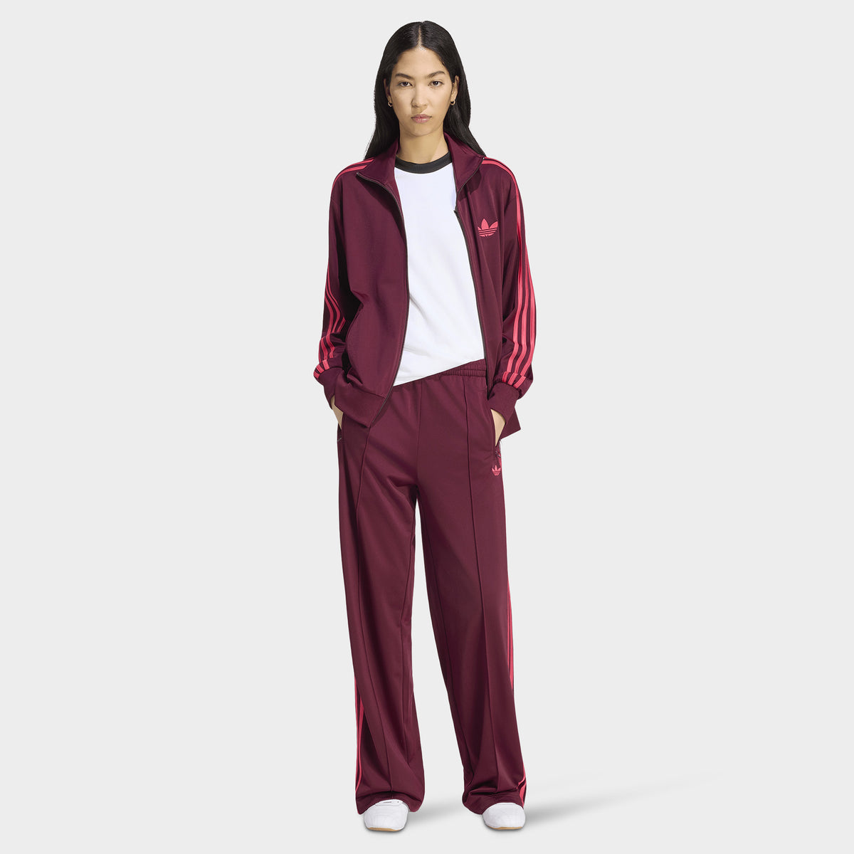 adidas Firebird Loose Track Pant. Full-body view of model wearing maroon track pants with power pink side stripes, styled with a white top and matching track jacket.