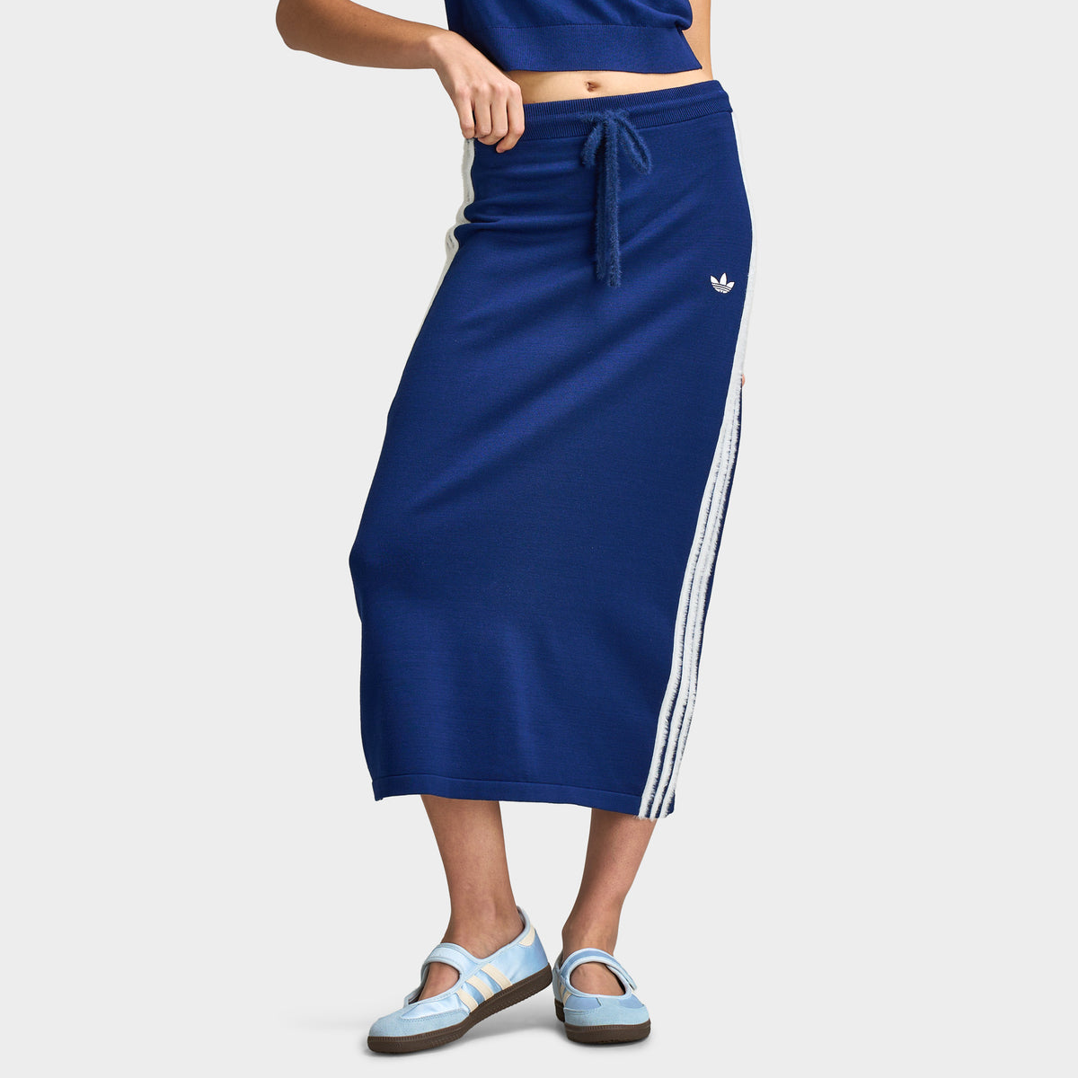 adidas 3 Stripe Knitted Skirt. Lower body front model view of a dark blue knitted midi skirt featuring an adjustable drawstring waistband, small white embroidered Trefoil logo on the left hip, contrast white 3-Stripes down the sides, and straight hem.