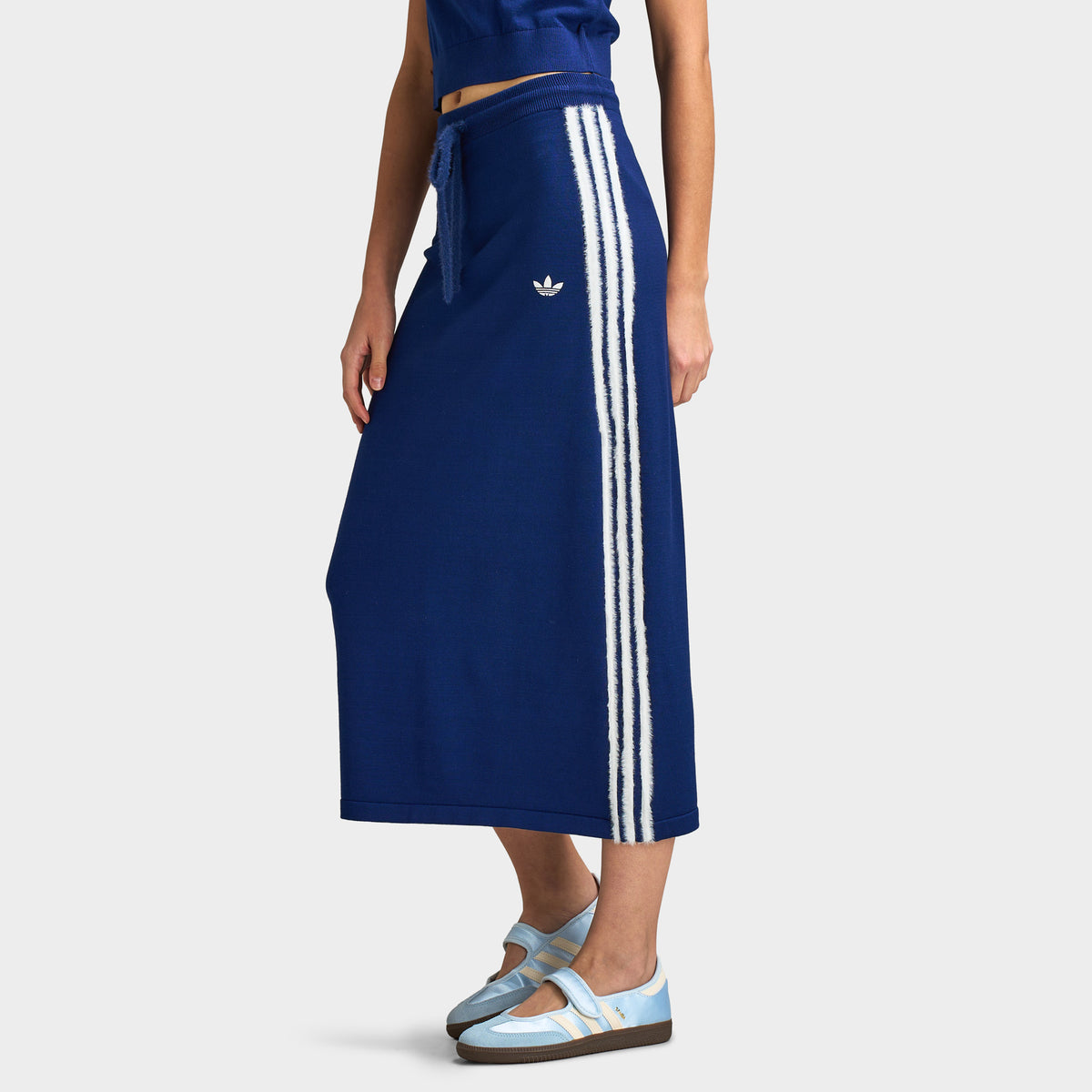 adidas 3 Stripe Knitted Skirt. Lower body three-quarter front model view showing the dark blue fine-knit midi skirt, white embroidered Trefoil logo, side seam white 3-Stripes, drawstring waist, and straight fit styled with light blue shoes. 
