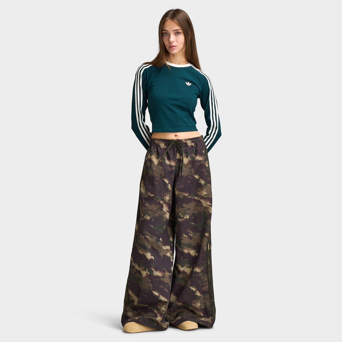 adidas Women's Camouflage Adilenium Track Pant. Full-length front model view of night cargo, aurora coffee, and olive strata camouflage wide-leg track pants, styled.  