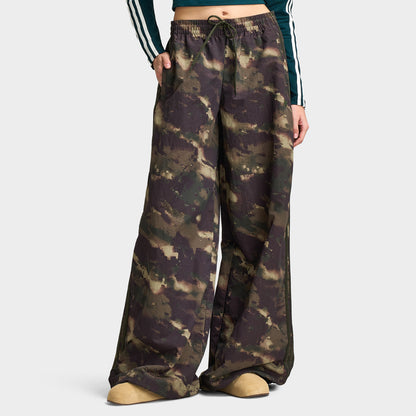adidas Women's Camouflage Adilenium Track Pant. Lower body front model view of night cargo, aurora coffee, and olive strata camouflage print wide-leg track pants featuring an elasticated waistband with adjustable drawstring, side pockets, tonal 3-Stripes along the side seams, wide-leg loose cut, and floor-length open hem.