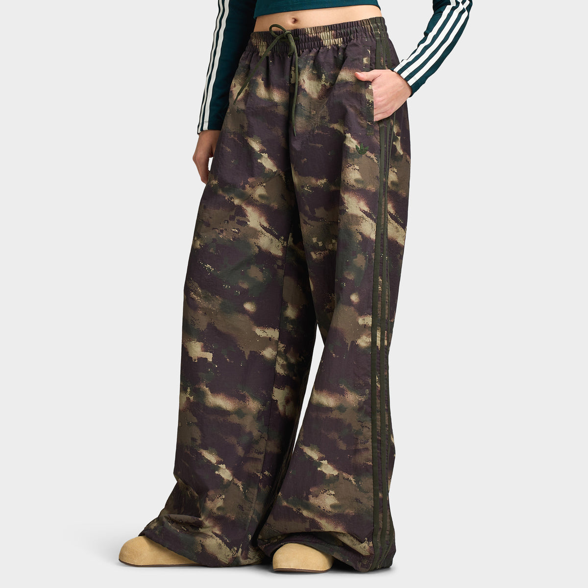 adidas Women's Camouflage Adilenium Track Pant. Three-quarter front model view displaying night cargo, aurora coffee, and olive strata camouflage track pants, elastic waistband, side pockets, wide-leg oversized fit, and open hems. 