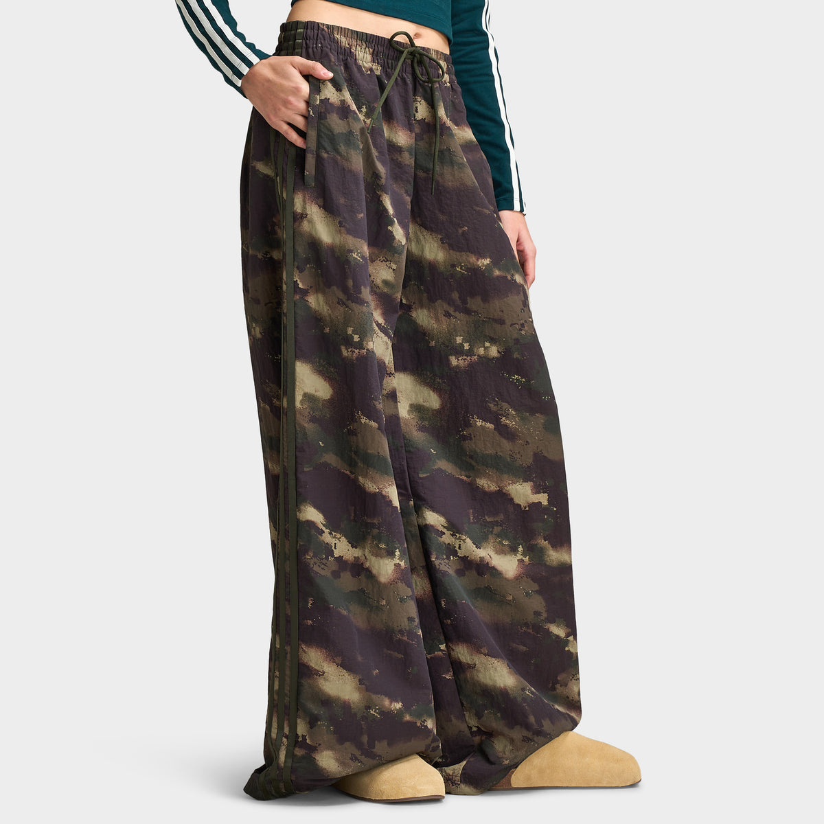 adidas Women's Camouflage Adilenium Track Pant. Side profile model view showing night cargo, aurora coffee, and olive strata camouflage track pants, side seam 3-Stripes detail, wide-leg cut, and open hem. 