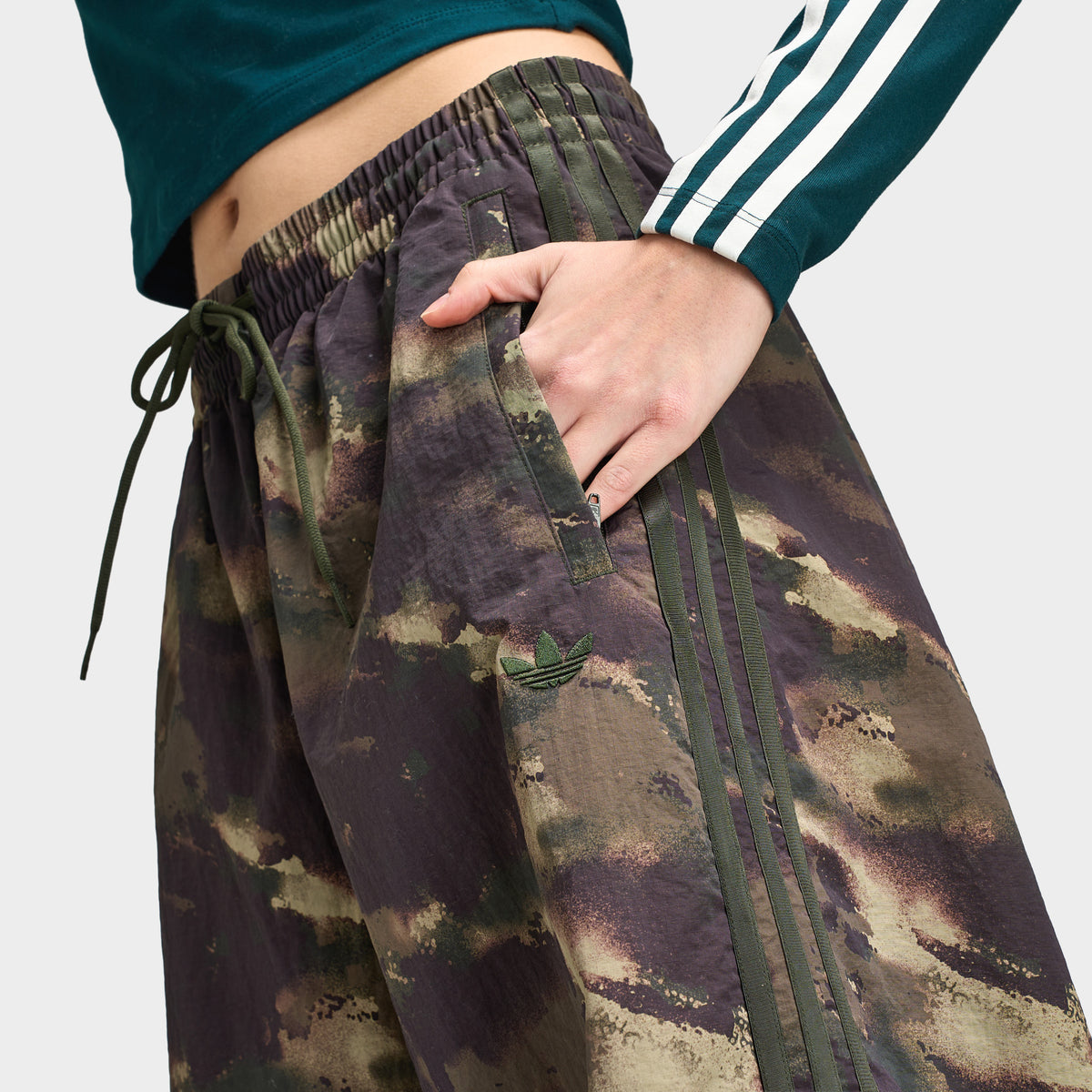 adidas Women's Camouflage Adilenium Track Pant. Close-up view of upper hip and waist highlighting night cargo, aurora coffee, and olive strata camouflage pattern, elasticated waistband with external drawstring, hand in side pocket, and tonal 3-Stripes side taping. 
