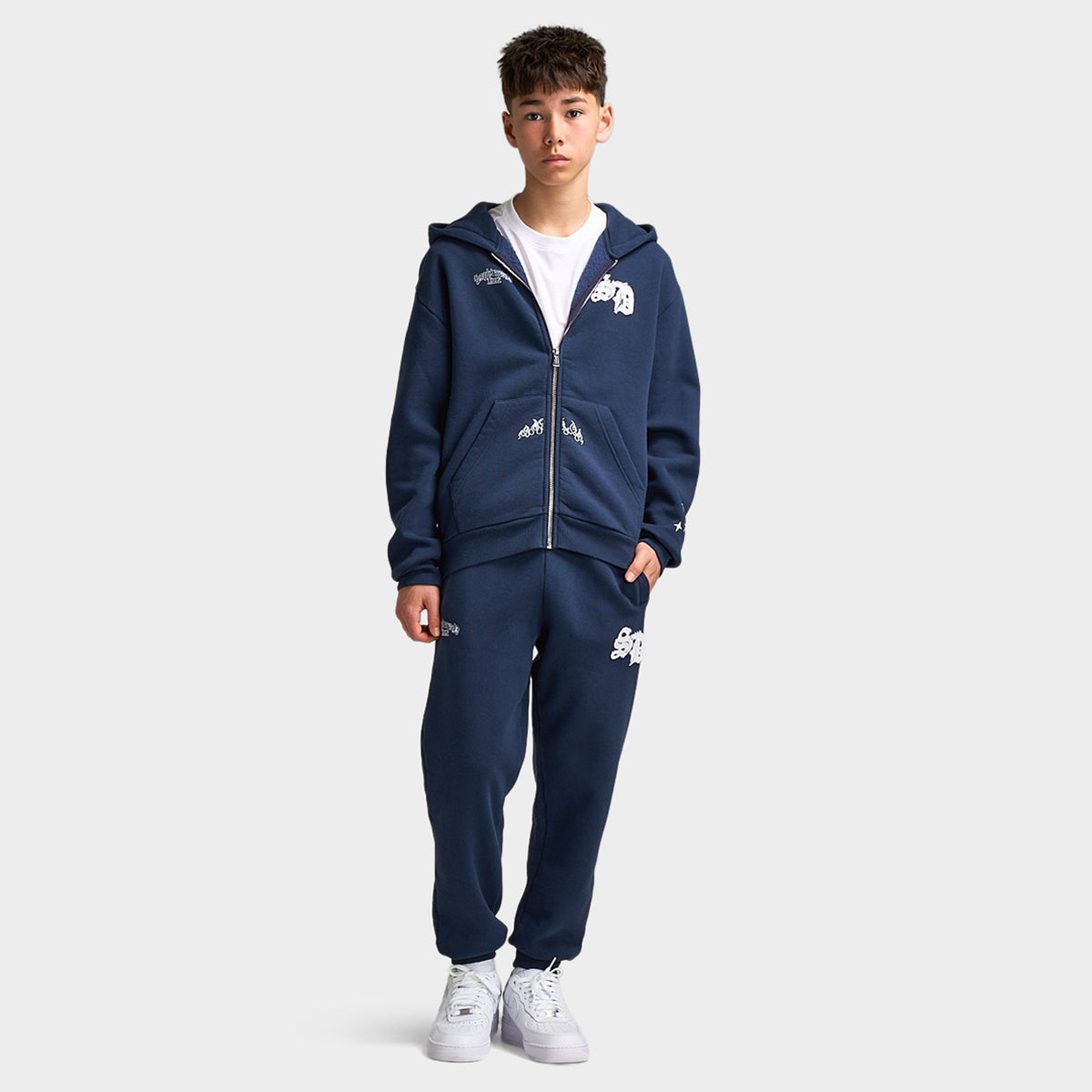 Supply & Demand Juniors' Scape Pants. Full-body view of model wearing washed navy pants with matching zip hoodie and white sneakers.