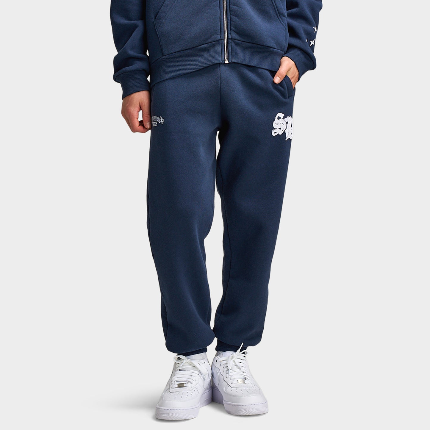 Supply & Demand Juniors' Scape Pants. Front view of model wearing washed navy pants with elastic waistband, tapered legs and white graphic branding.