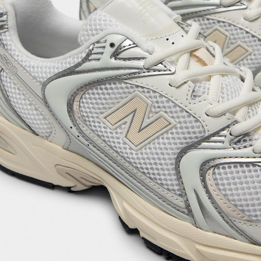 New Balance 530. Close-up of silver metallic and linen sneaker showing cream laces, mesh panels, metallic overlays and N logo.