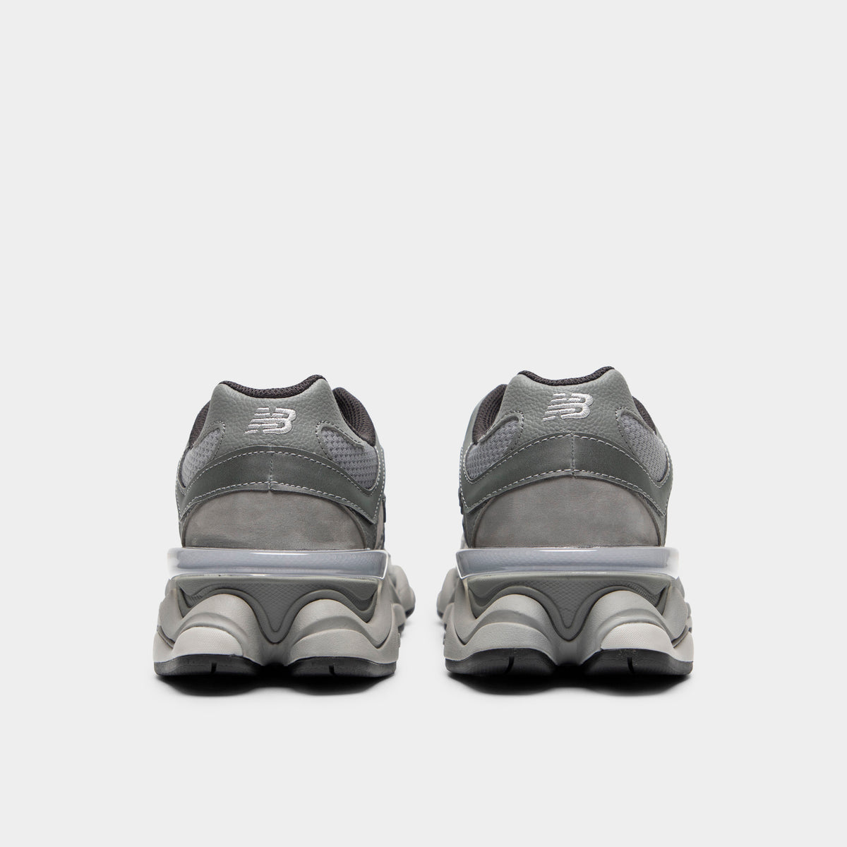 New Balance 9060. Rear view of a pair of chunky sneakers displaying slate grey suede heel counters with silver "NB" embroidery, translucent CR heel clips, dark grey padded collar linings, and exaggerated sculpted pod midsoles. 