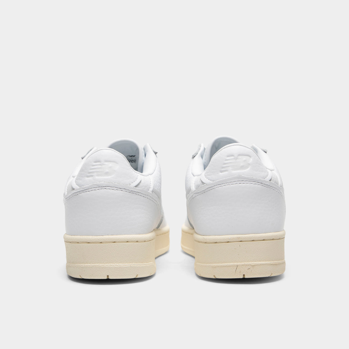 New Balance 950. Rear view of a pair of low-top sneakers displaying white leather heel counters with debossed "NB" logos, white padded collars, and off-white aged rubber midsoles. 