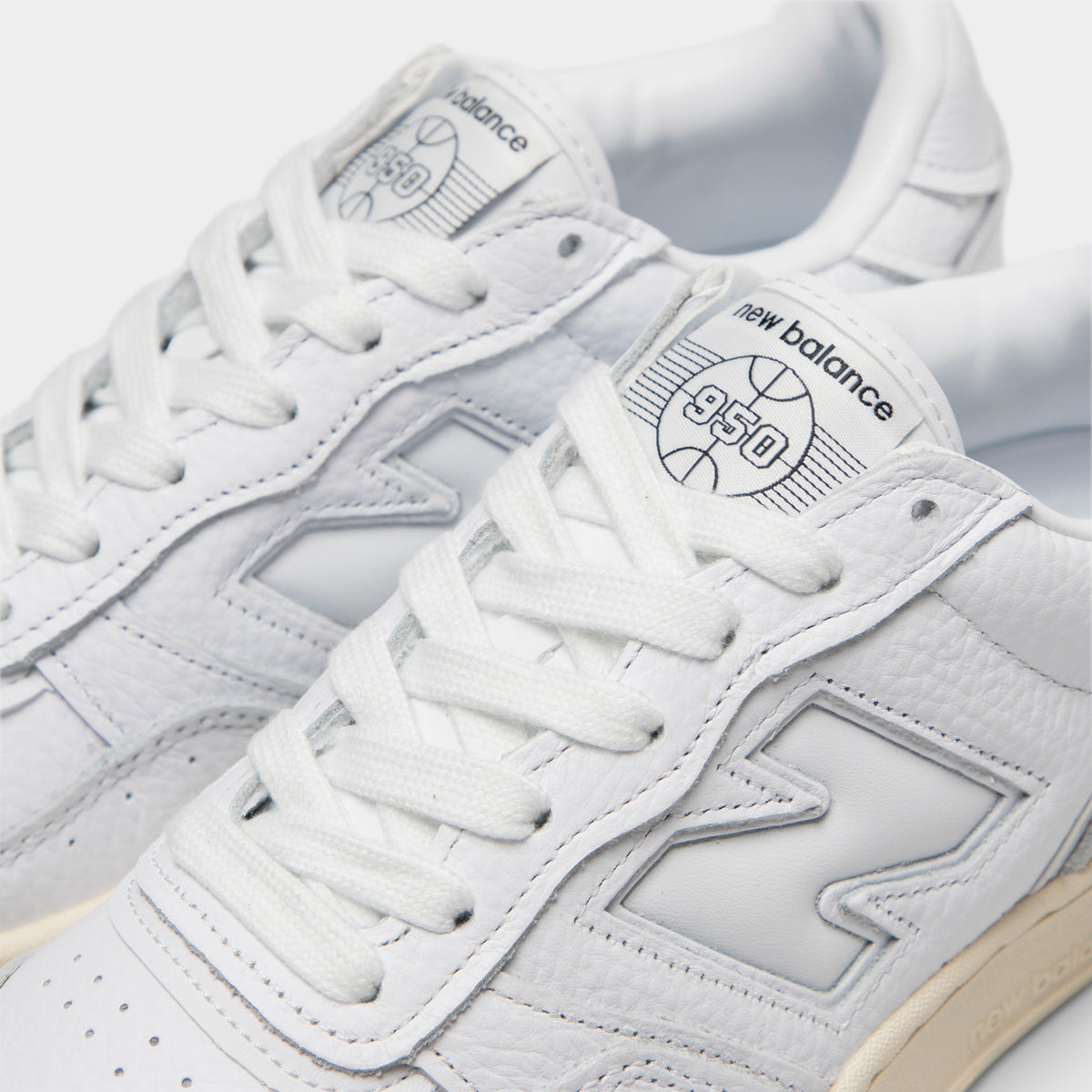 New Balance 950. Close-up view of tongue and lacing highlighting white leather construction, white "N" midfoot logo, white laces, and white leather tongue patch with grey New Balance 950 basketball graphic print. 
