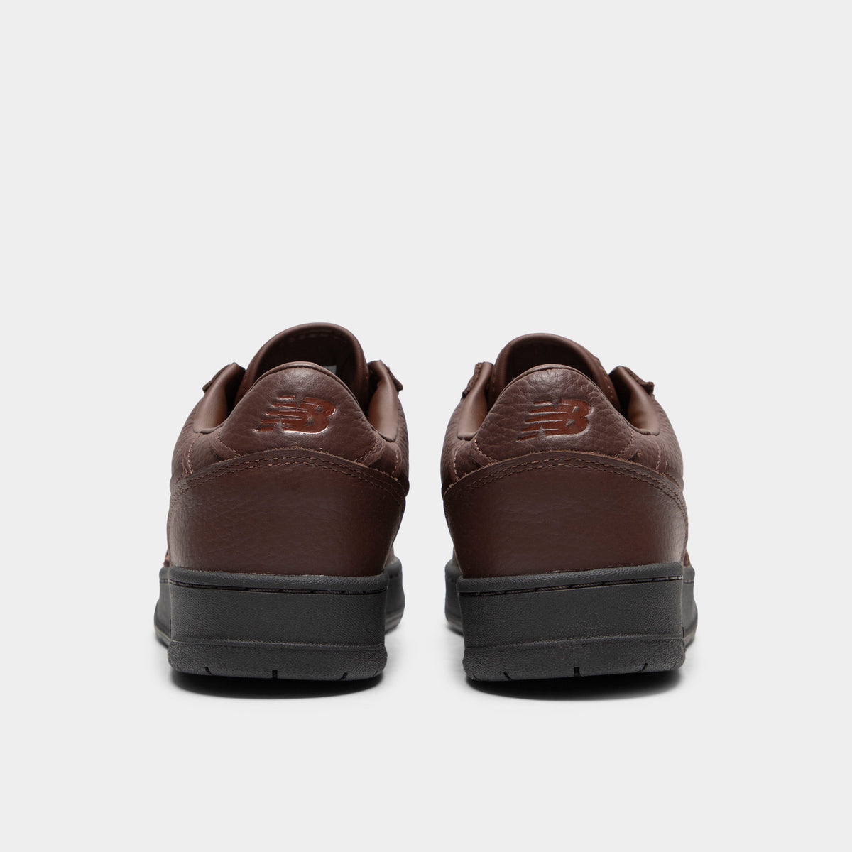 New Balance 950. Rear view of a pair of low-top sneakers displaying dark brown tumble leather heel counters with debossed "NB" logos, dark brown padded collars, and black rubber midsoles. 