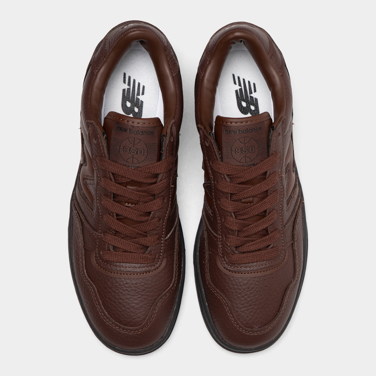 New Balance 950. Top-down overhead view of a pair of low-top sneakers displaying dark brown tumble leather toe boxes, dark brown laces, leather tongues with debossed logo patches, and white insoles with dark "NB" branding. 