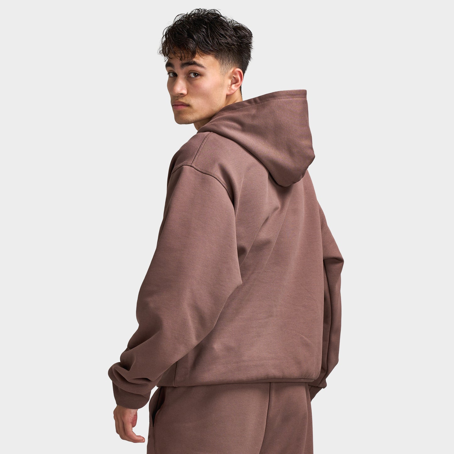 Unlike Humans Ridge Hoodie. Back three-quarter view of model wearing coffee quarts hoodie with raised hood and loose silhouette.